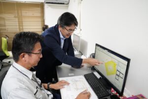 事務所でパソコンに表示した測量図を指しながらスタッフと打ち合わせをする土地家屋調査士